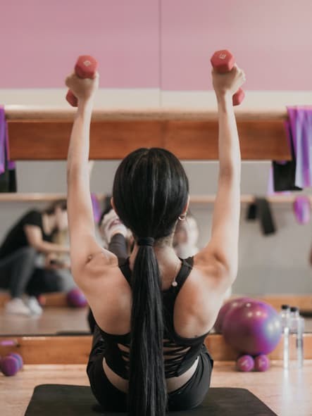 back of person, in gym arms raised holding dumb bells